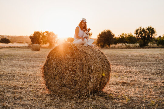 Smiling Woman With Jack Russell Terrier Sitting Cross-legged On Straw Bale During Sunset