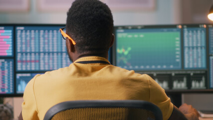 Rack focus of black man in yellow t shirt analyzing financial graphs on computers while trading on stock exchange market during work from home