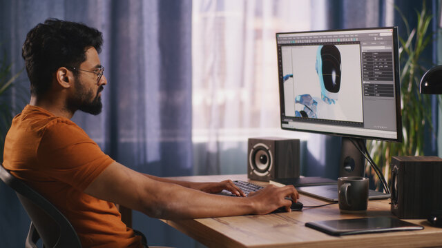 Indian male developer using computer to create and render 3D model of futuristic robot on computer during work in dim living room in evening