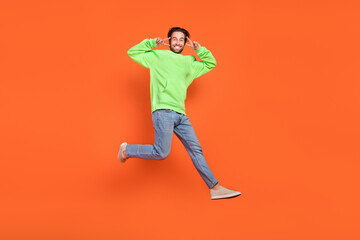 Full body photo of brunet millennial guy run show v-sign wear hoodie jeans footwear isolated on orange background