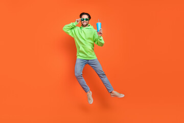 Fototapeta na wymiar Full size photo of brunet young guy drink wear sweatshirt jeans sneakers eyewear isolated on orange background