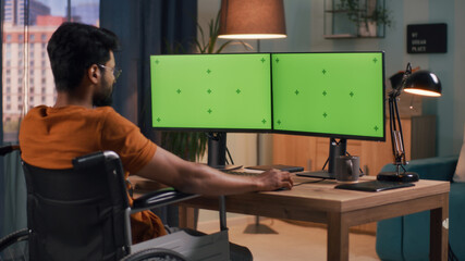 Indian male sitting on wheelchair and browsing computer with chromakey monitors in evening at home