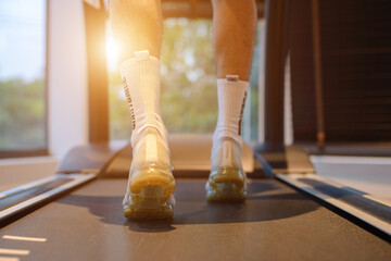 Young athlete wearing white sport shoes running on treadmill in the morning with sunlight. He training for cardiovascular endurance and good health with personal trainer in fitness center.