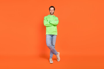 Fototapeta na wymiar Full size photo of brunet young man stand wear green hoodie jeans sneakers isolated on orange color background