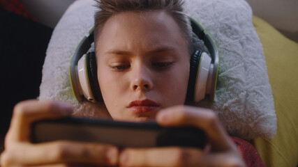 From above male teenager in wireless headphones lying on bed and playing videogame on cellphone at home