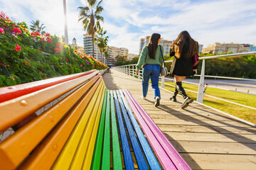 Two young women walking by a bench painted with rainbow colours