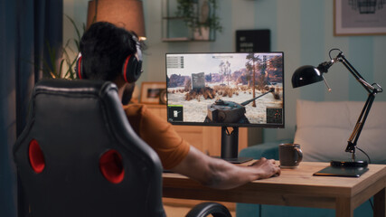 Back view of guy in casual clothes and headphones playing tank videogame on computer while sitting...