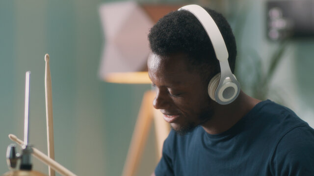 Young African American man listening to rhythmic music in wireless headphones and playing drums during rehearsal in home studio in daytime