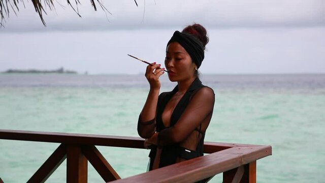 a girl in a black evening dress in a retro style stands at a water villa and smokes a cigarette through a mouthpiece against a background of blue sky and azure sea. Maldives resort.