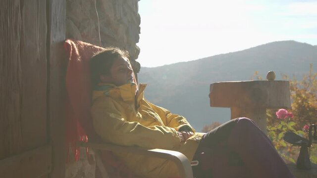 Overview of young girl in yellow jacket, sitting outside cabin chilling after hiking. Mountains on the background. Sunny autumn day.