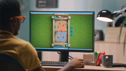 African American male gamer playing videogame on computer while sitting at table in evening at workplace