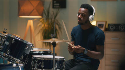 Fototapeta na wymiar Black man in wireless headphones playing rhythmic tune on drums during rehearsal in home studio in evening