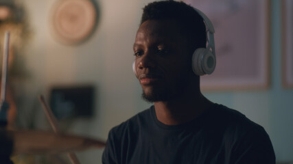 African American man in wireless headphones enjoying process of playing drums during rehearsal in evening at home