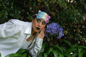 Young latin disguised woman make up and colorful mask over her head listening a blue flower on natural background