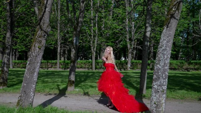 a blonde with big breasts and in a lush red dress runs along the path of the park between the trees. side view. the camera moves in parallel