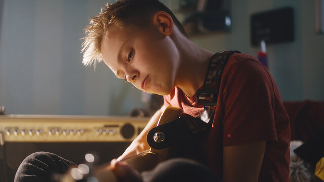 Focused teen musician playing electric guitar and adjusting controls on amplifier while sitting on bed during music session in sunlit bedroom at home