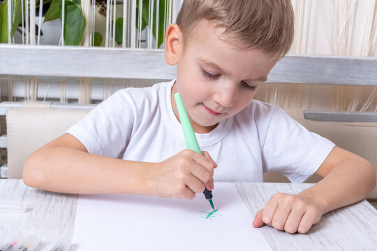 A lovely emotional little boy draws a green Christmas tree with a felt-tip pen, pencil on a sheet of paper while sitting at the table