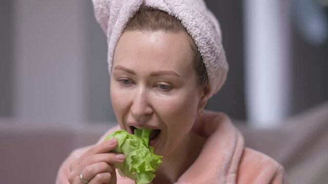 Tired dissatisfied young woman in hair towel eating green salad leaf sighing. Portrait of unhappy Caucasian millennial lady dieting at home in the morning. Femininity and self-discipline concept