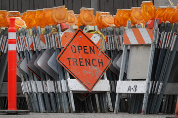 Orange and yellow traffic control barricades, topped with flashing lights, are shown with an OPEN TRENCH sign and a delineator pylon cone in preparation for a road infrastructure project.