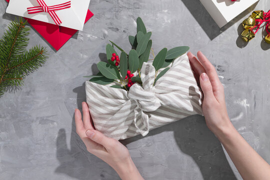 Female hands wrapping a japanese furoshiki style gift with eucalyptus and red berries on a gray background