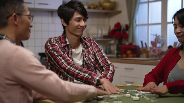 selective focus with slow motion asian son in red plaid shirt looking at other family players with cunning smile while shuffling tiles, feeling confident that he will win the mahjong game