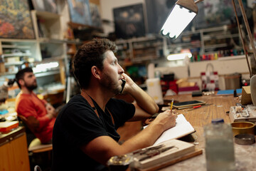 Young man in his craft workshop talking on the phone, picking up orders and writing down a notebook.