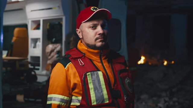 Male paramedic in uniform and hat looking at camera while standing near ambulance car during rescue mission after disaster at night