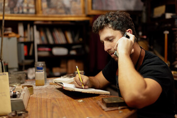 Young man in his craft workshop talking on the phone, picking up orders and writing down a notebook.