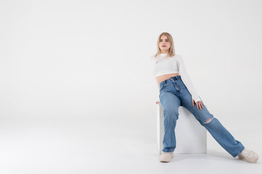A young girl in a white sweater and jeans sits and poses on an isolated white background in the studio. People lifestyle concept. Copy space for copy