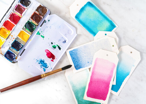 Open watercolor paint box with brush painting gift tags.