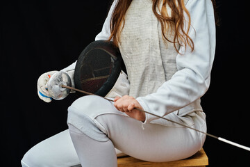 Close up of a fencer dressed in white fencing, holding his mask and a sword on black background.