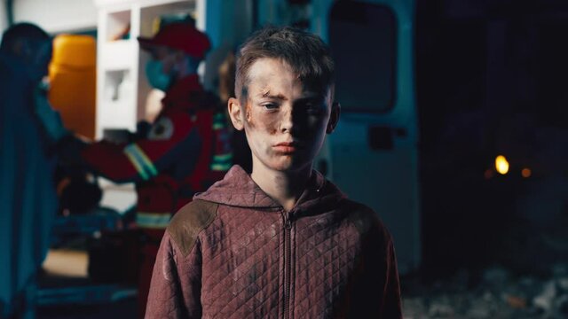 Teenager with dirty face looking at camera while standing near ambulance van after accident at night