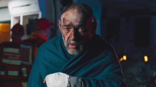 Concussed elderly man wrapped in blanket looking at camera while standing near paramedic van after accident at night
