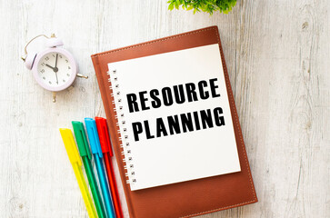 Notepad with the text RESOURCE PLANNING on a wooden table. Brown diary and pens.