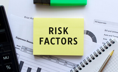 Text RISK FACTORS on the page of a notepad lying on financial charts on the office desk.