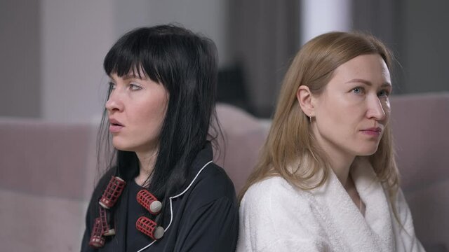 Portrait of two dissatisfied women sitting indoors sighing rolling eyes. Argued displeased Caucasian female adult friends with sarcastic facial expression in living room at home. Slow motion