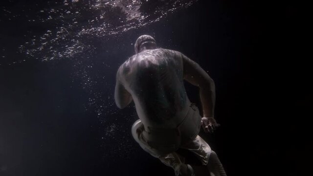 a large bald man without a shirt hovers in the dark under the water in the pose of a baby