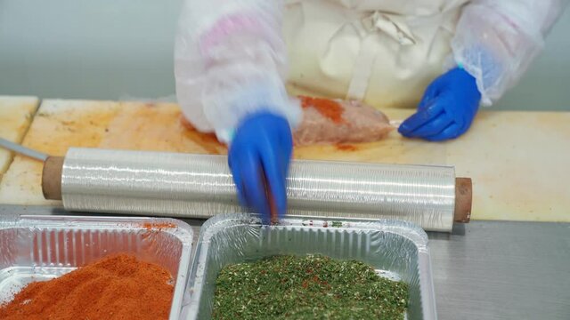 Front motion of young female butcher processing sausage at meat factory, adiing colorful spice. Worker wearing white uniform making meat product in traditional way, rolling using clingfilm on table.