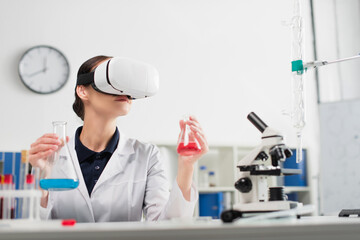 Scientist in vr headset holding flasks while working near microscope and test tubes in laboratory.