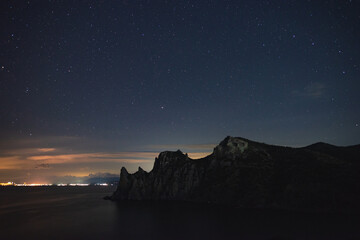 A large panorama of a beautiful view of the sea among the mountains and a beautiful starry sky with the Milky Way. The city by the sea glows at night in the distance
