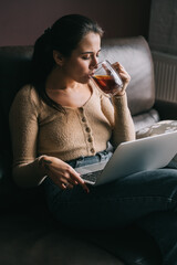 Young woman work at home or stydy on her laptop, holding cup of coffee or tee. Freelance concept. Female sit on leather couch at home.