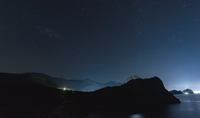 A large panorama of a beautiful view of the sea among the mountains and a beautiful starry sky with the Milky Way. The city by the sea glows at night in the distance