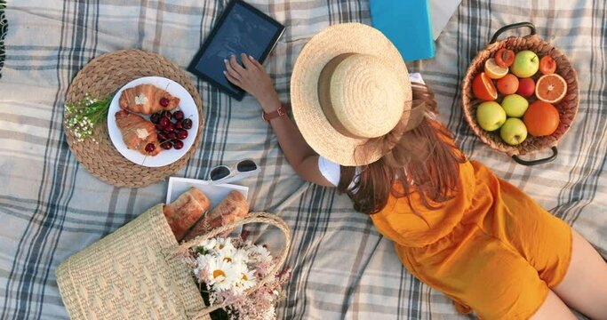 Top view of young beautiful woman in hat lying on picnic in park and looking at photos on tablet, enjoying weekend.