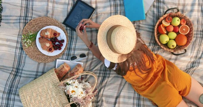 Top view of young beautiful stylish woman in hat lying on picnic in park and enjoying ripe cherries watching movie on tablet.