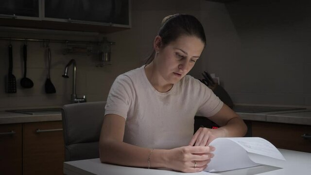 Confused woman examines financial documents on kitchen at night. Concept of financial difficulties, bankruptcy, taxes and rent payment.