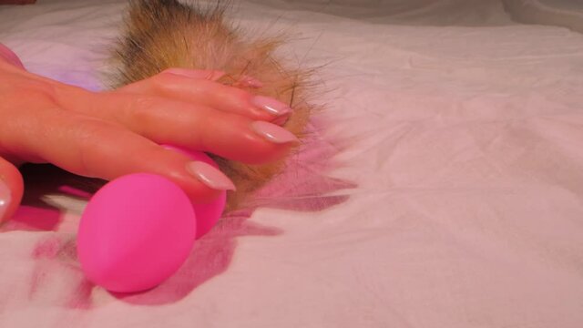 Woman hand puts colorful anal plug with fluffy tail on bed with sheet and light becomes dark purple in semi-dark room closeup
