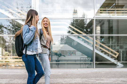 Two Female College Classmates Walking While Chatting With A Glass Building In The Background.