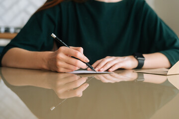 Close-up hands of unrecognizable young woman writing handwritten letter sitting at table at home, selective focus, blurred background. Happy female writing wishes on Christmas card.