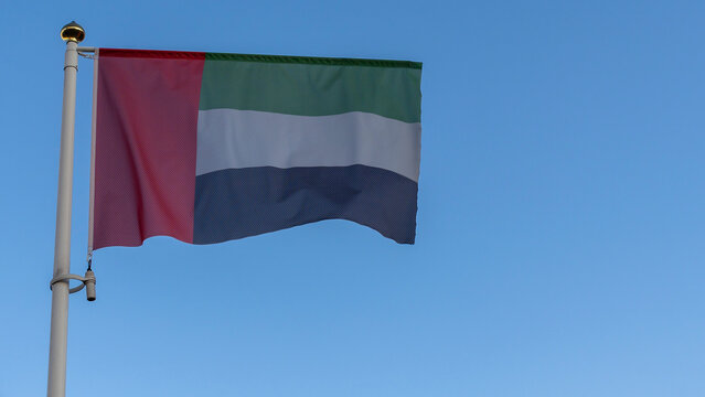 National flag of United Arab Emirates on a flagpole in front of blue sky with sun rays and lens flare.