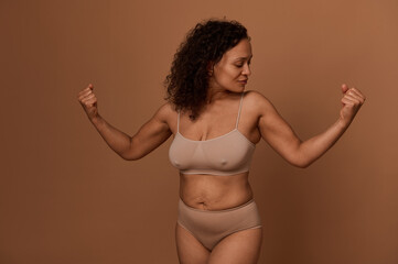 Delighted confident middle aged African American woman with stretch marks, in beige underwear looking at her arms and clenched fists, posing against colored background with copy space.